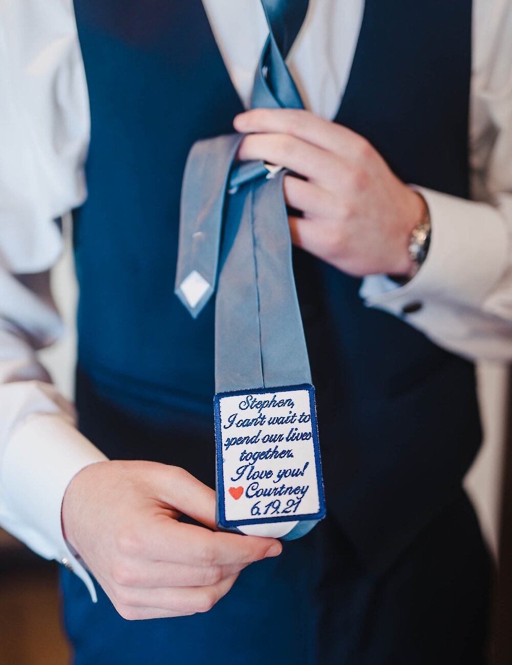 Close-up of the groom holding his tie, revealing a personalized label embroidered with a blue border, a heart, and a love note from Courtney.