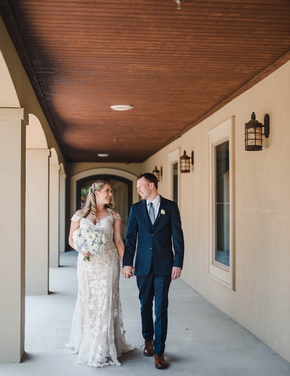 The bride in a lace gown and the groom in a navy suit walk hand-in-hand and smile at each other down a covered outdoor hallway with a wooden ceiling.