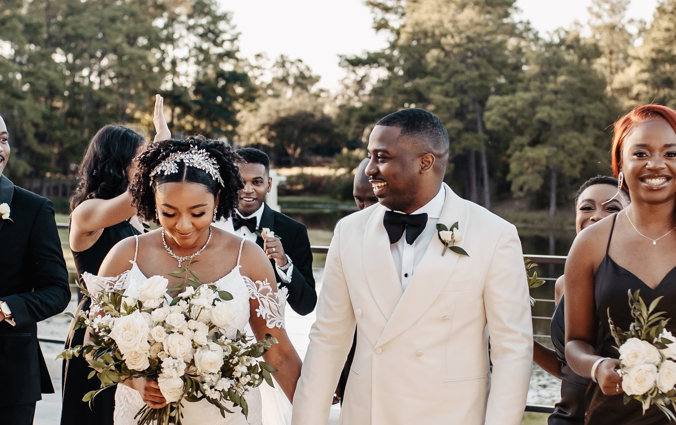The smiling bride and groom walk hand-in-hand during their recessional, with the groom in a white tuxedo jacket and the bride holding a large white rose bouquet.