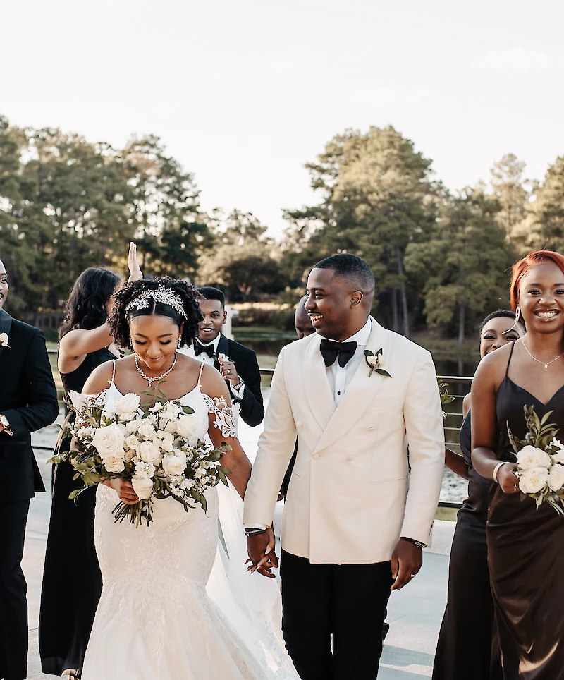 The smiling bride and groom walk hand-in-hand during their recessional, with the groom in a white tuxedo jacket and the bride holding a large white rose bouquet.