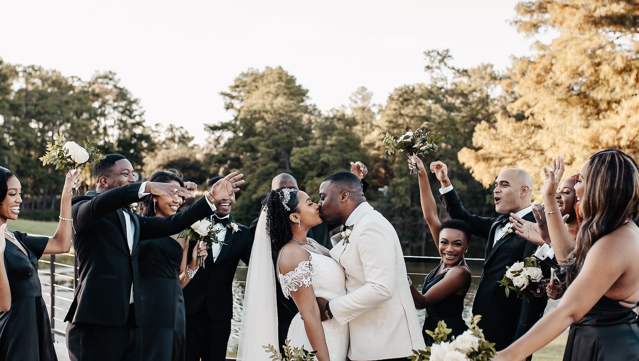 The bride and groom kiss while surrounded by their cheering wedding party, who are all dressed in black and white formal attire outdoors.