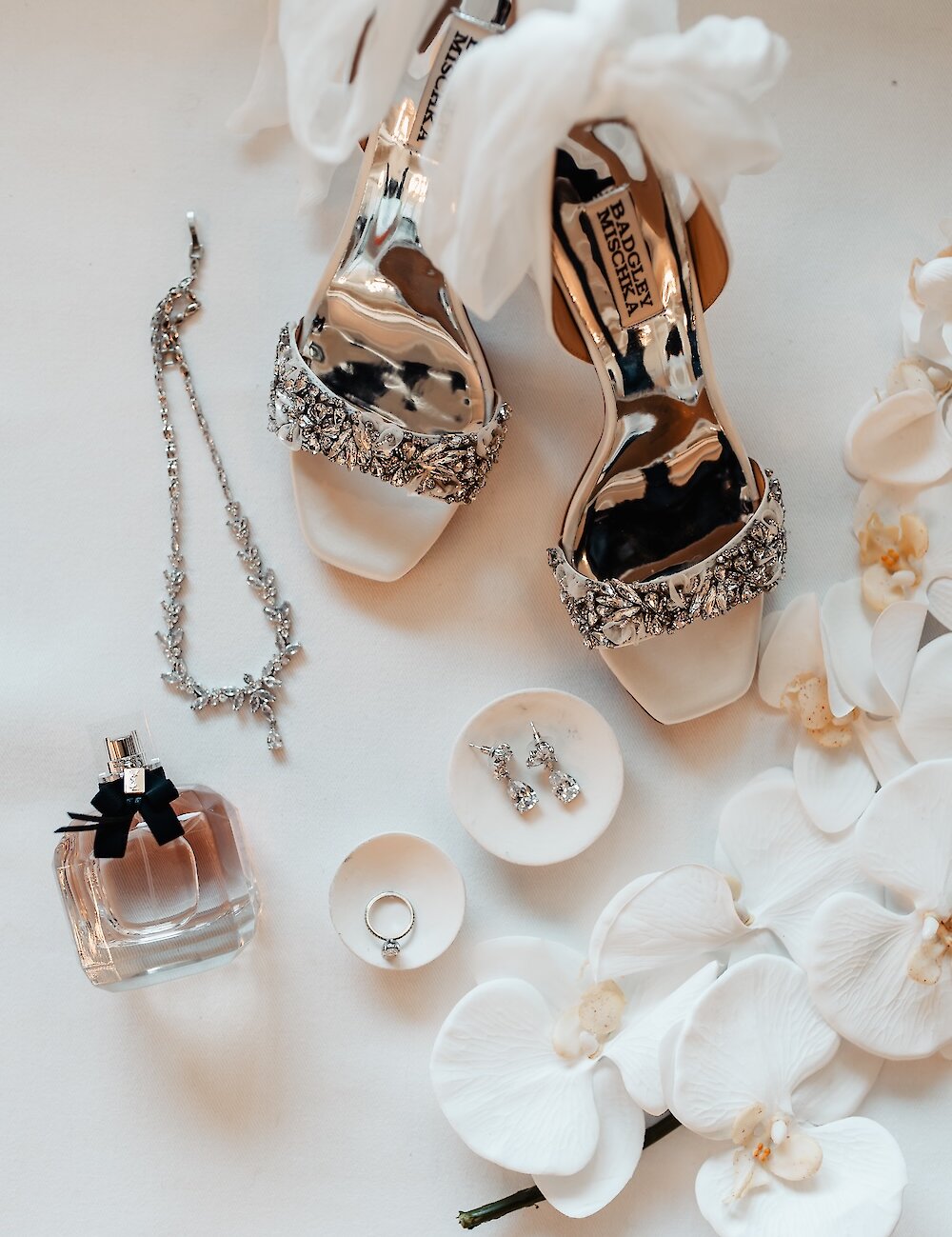 A wedding flat lay featuring silver jeweled Badgley Mischka bridal heels, a diamond necklace, earrings, engagement ring, and a square perfume bottle, surrounded by white orchid petals.