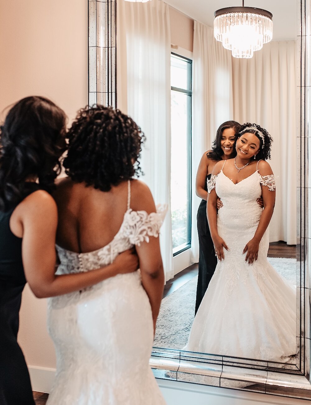 A smiling bride in her fitted lace gown is pictured in a large mirror, with a bridesmaid standing behind her, adjusting the dress in the bridal suite.