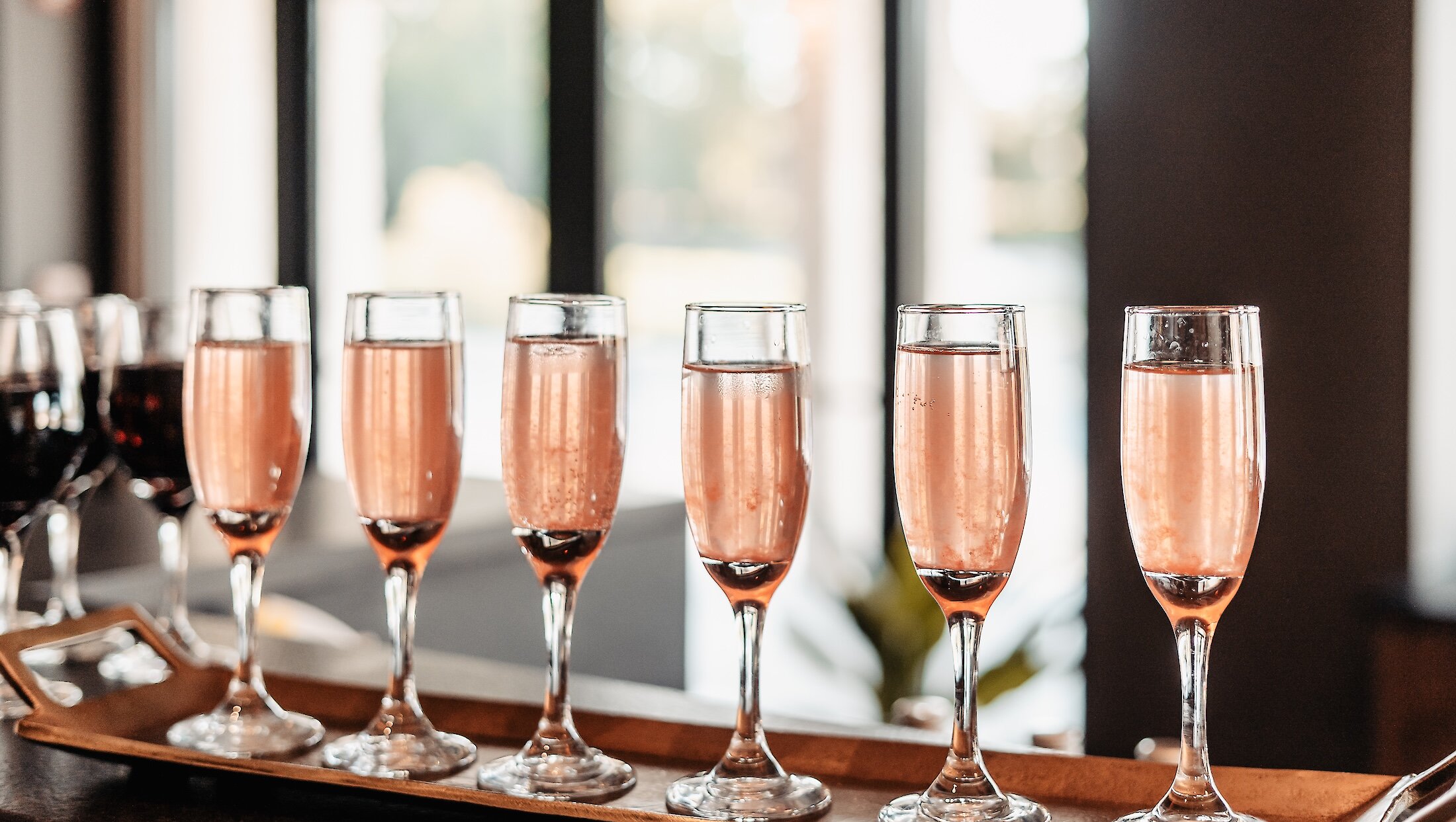 A close-up of a row of six champagne flutes filled with pink sparkling wine, displayed on a long golden tray at a reception bar.