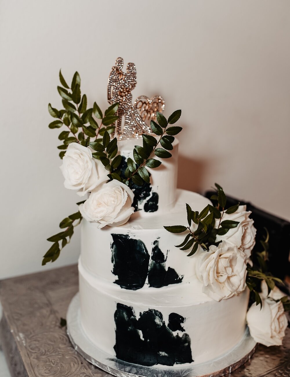 A two-tiered white wedding cake with black watercolor detailing, topped with a glittering topper and decorated with white roses and green leaves.