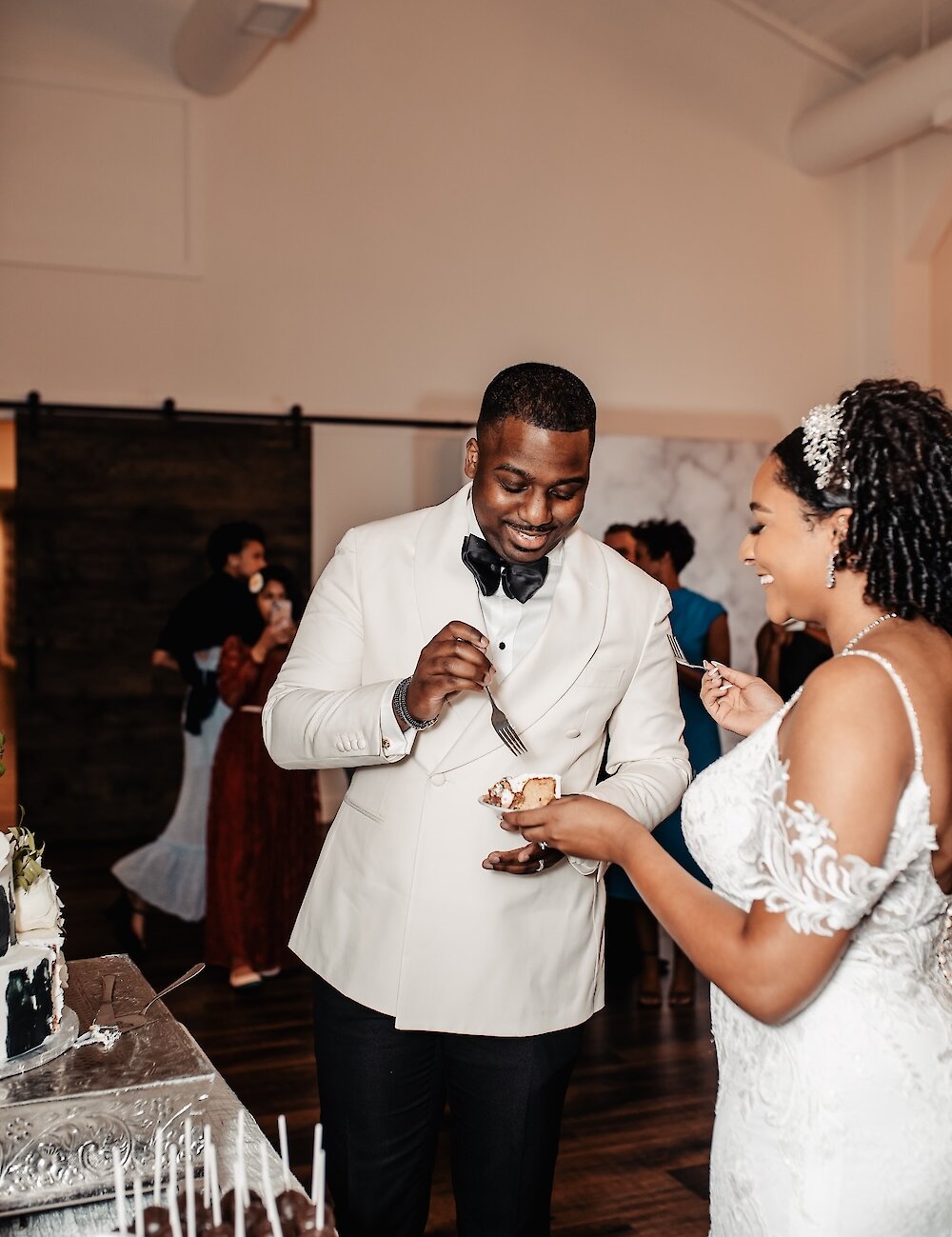 The smiling groom in a white tuxedo feeds a piece of wedding cake to his bride at the dessert table during the reception celebration.