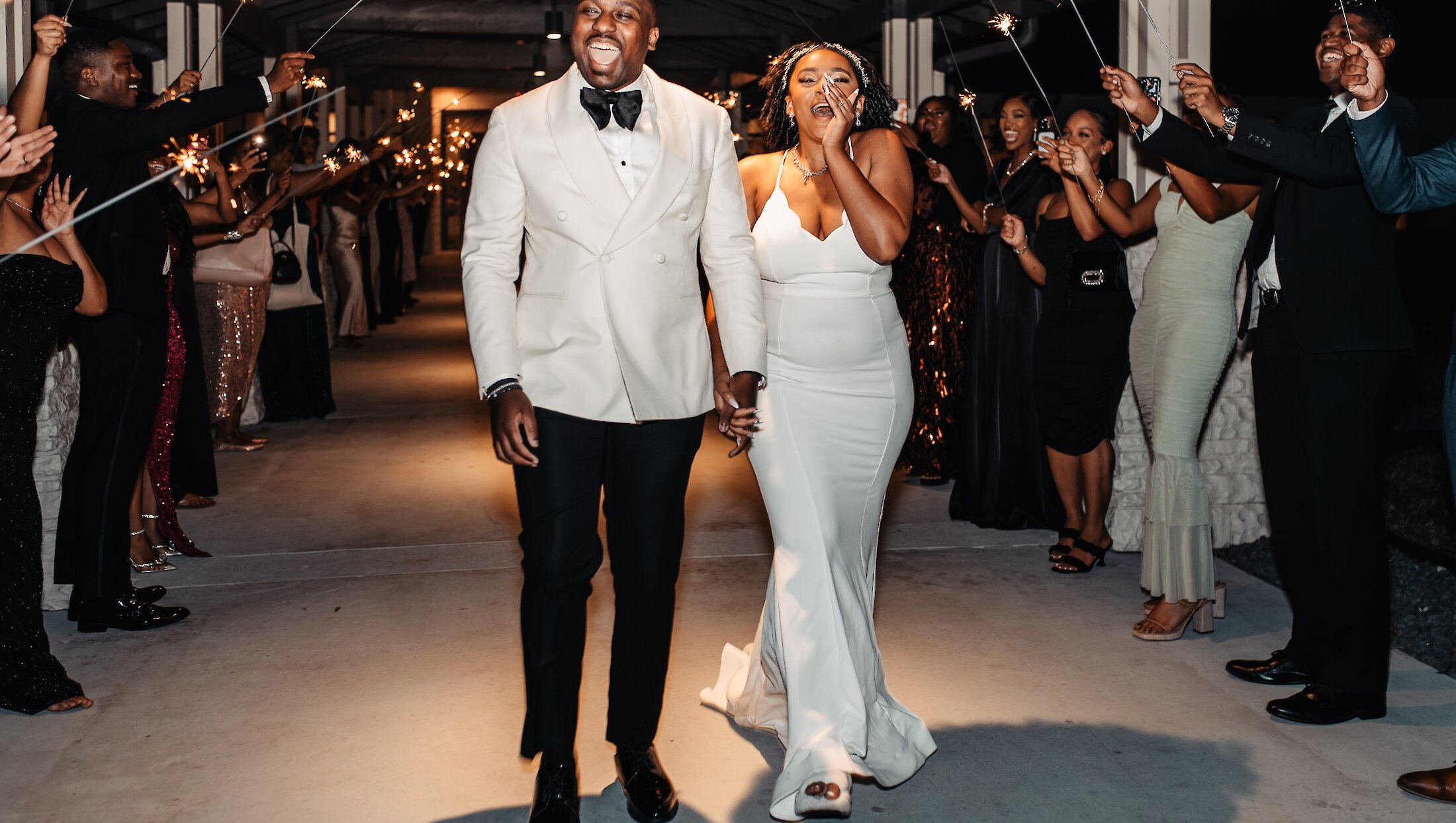 The elated bride and groom walk hand-in-hand through a celebratory tunnel of guests holding sparklers during their wedding exit.