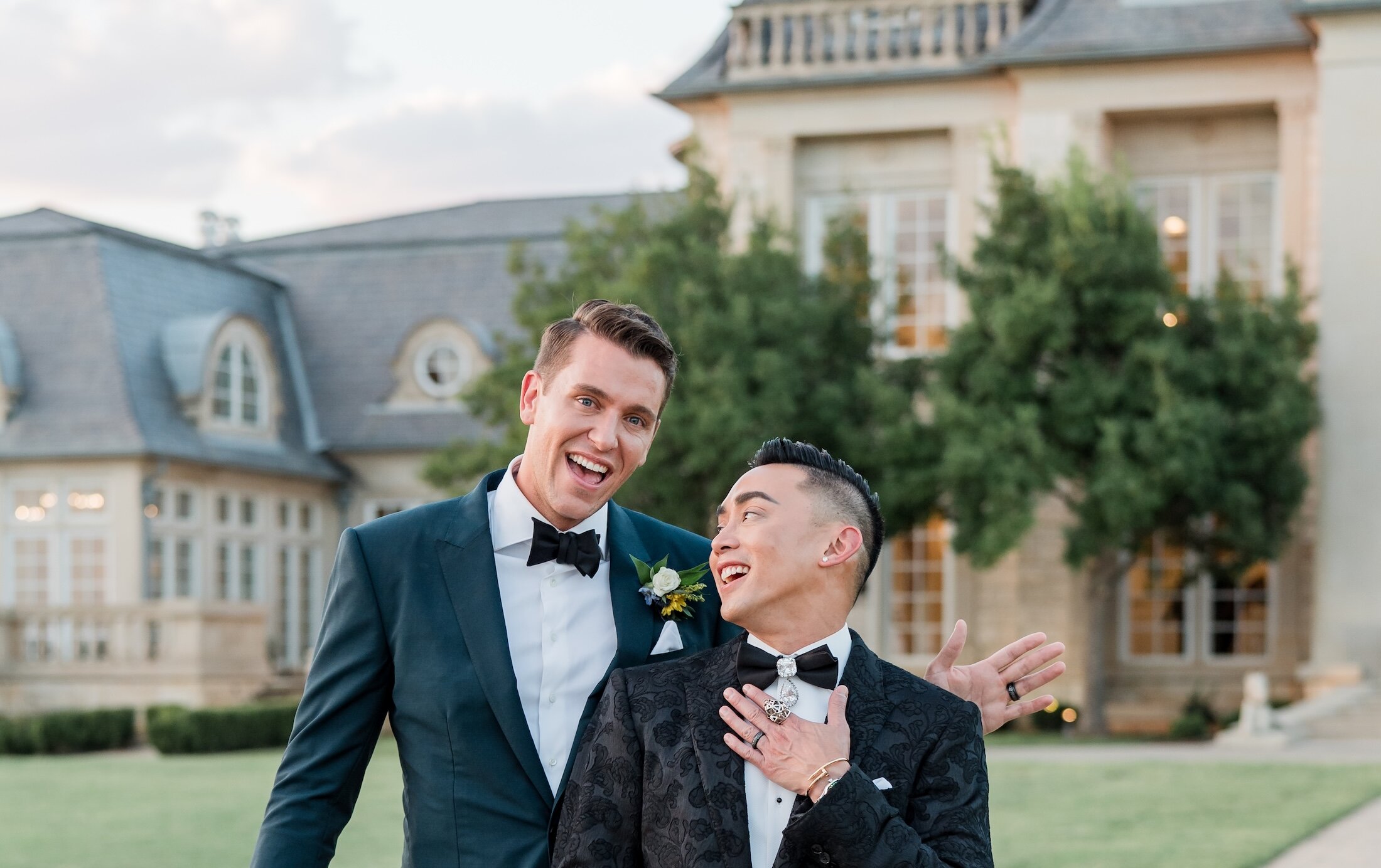Two joyful groomsmen pose laughing in front of a grand mansion venue, with one in a navy suit and the other in a black patterned tuxedo.