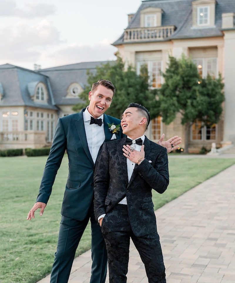 Two joyful groomsmen pose laughing in front of a grand mansion venue, with one in a navy suit and the other in a black patterned tuxedo.
