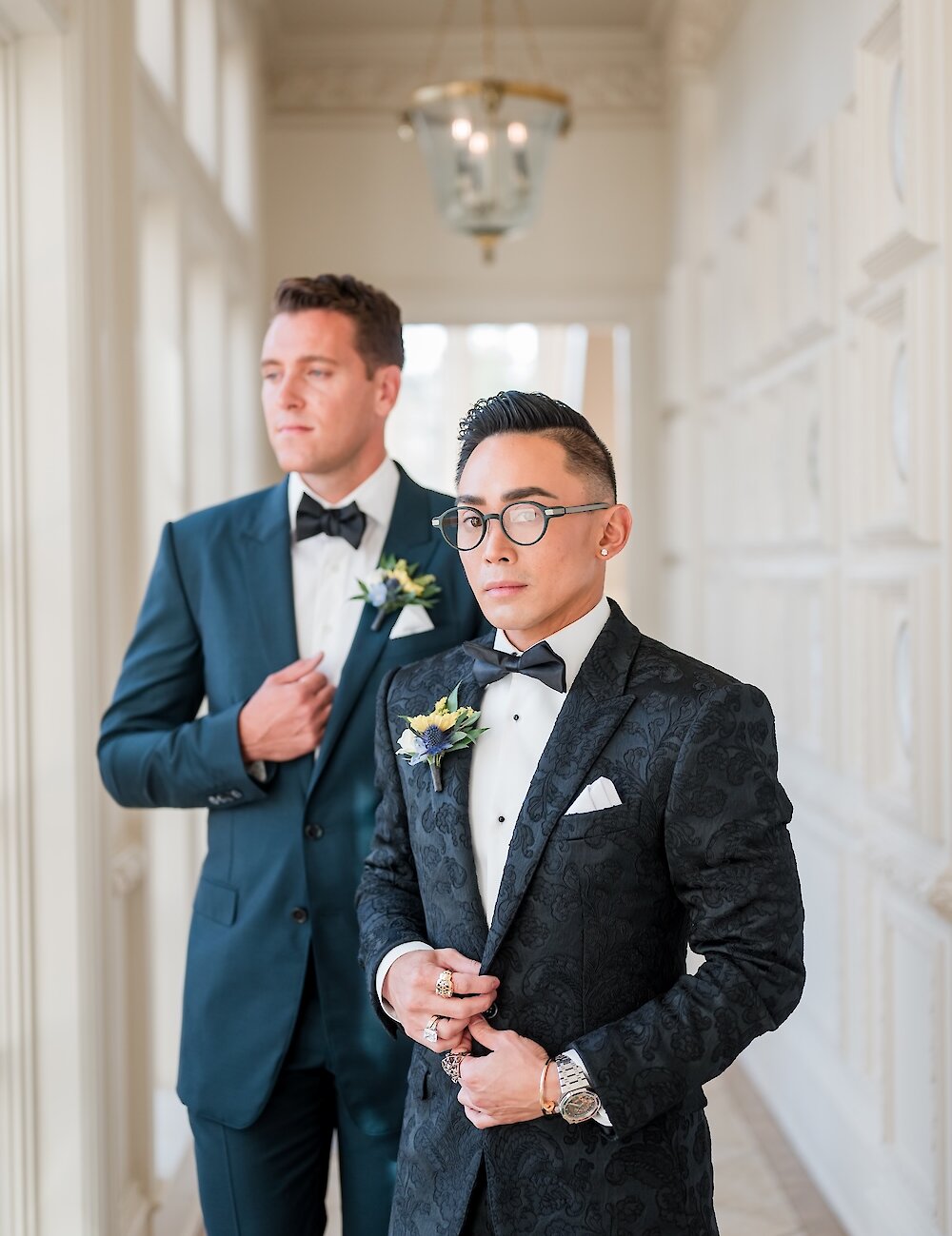 A formal portrait of the two grooms standing in a brightly lit hallway, with one in a navy suit and the other in a black floral-patterned tuxedo.