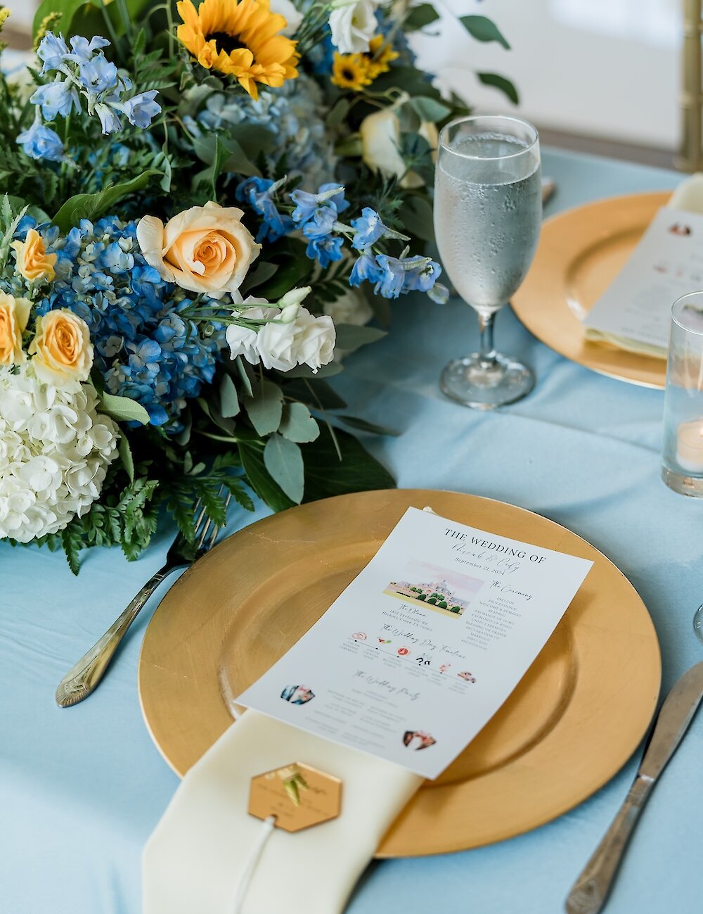 A reception place setting with a gold charger plate, a custom printed wedding program on a napkin, and a centerpiece featuring sunflowers and blue delphiniums on a light blue tablecloth.