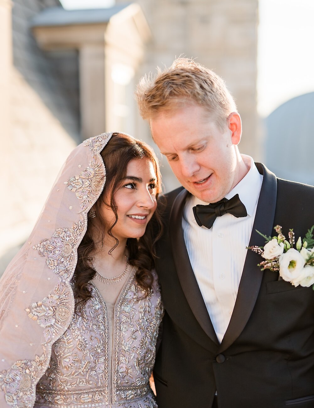 A romantic close-up of the bride in a lavender embroidered traditional gown and veil looking up at the groom in a classic black tuxedo with a white boutonniere.