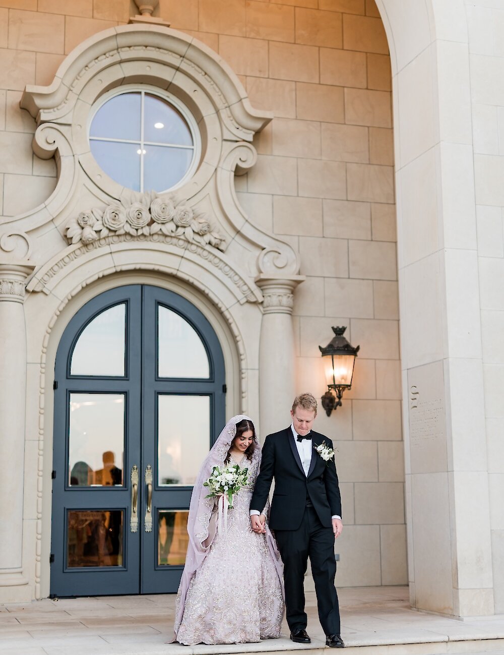 The bride in a long lavender traditional gown and the groom in a tuxedo walk down stone steps hand-in-hand outside a grand mansion entrance with carved detailing.