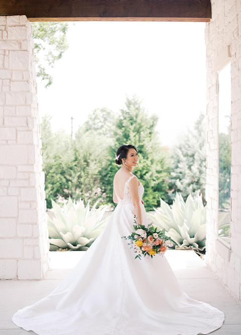 Bride in wedding dress with back deep v and bouquet outside stone exterior columns