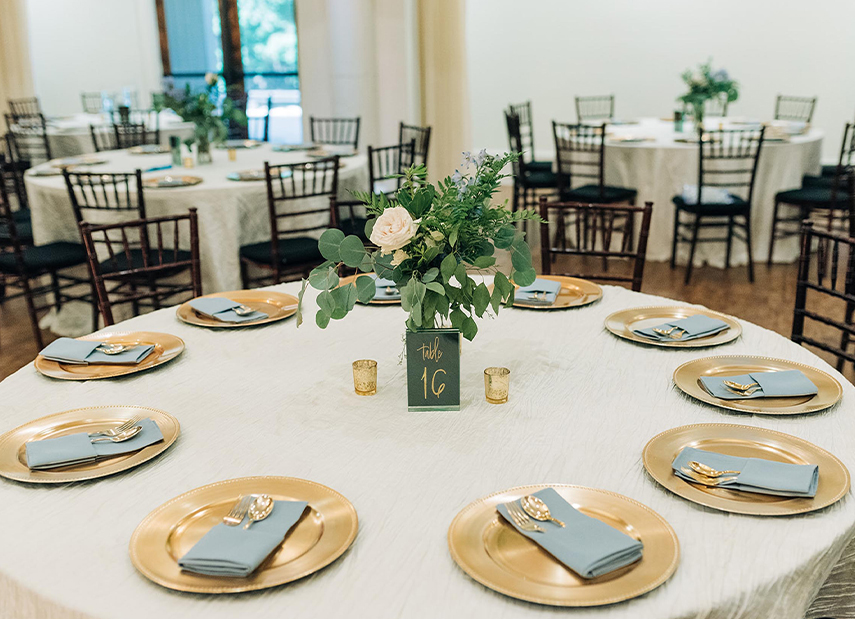 Top down view of set table with gold chargers and light blue napkins and simple centerpiece with a white rose