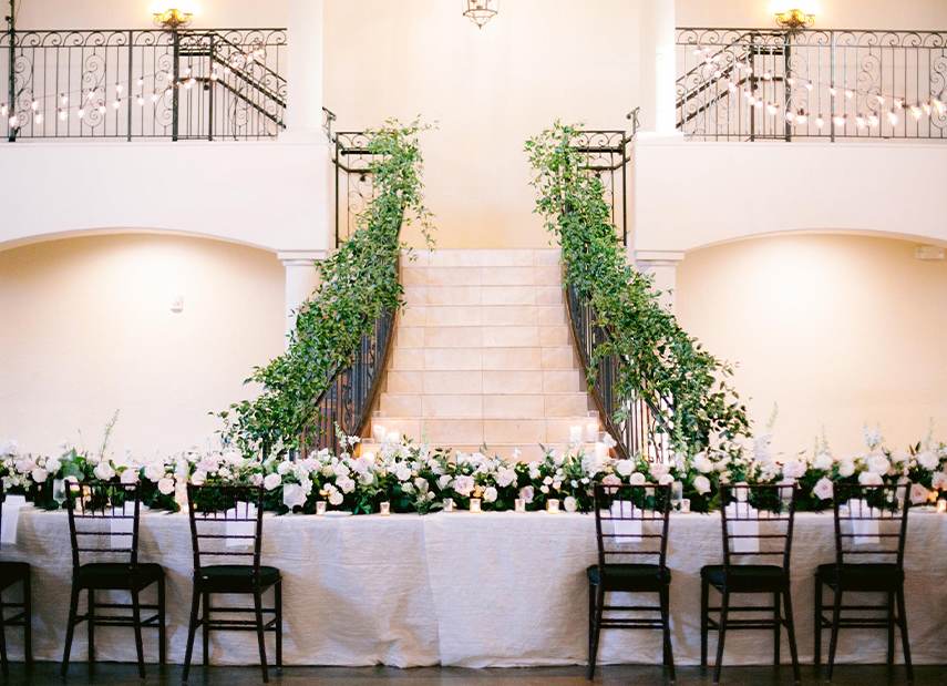 Long wedding party table with white linens and dark chairs, grand staircase in background