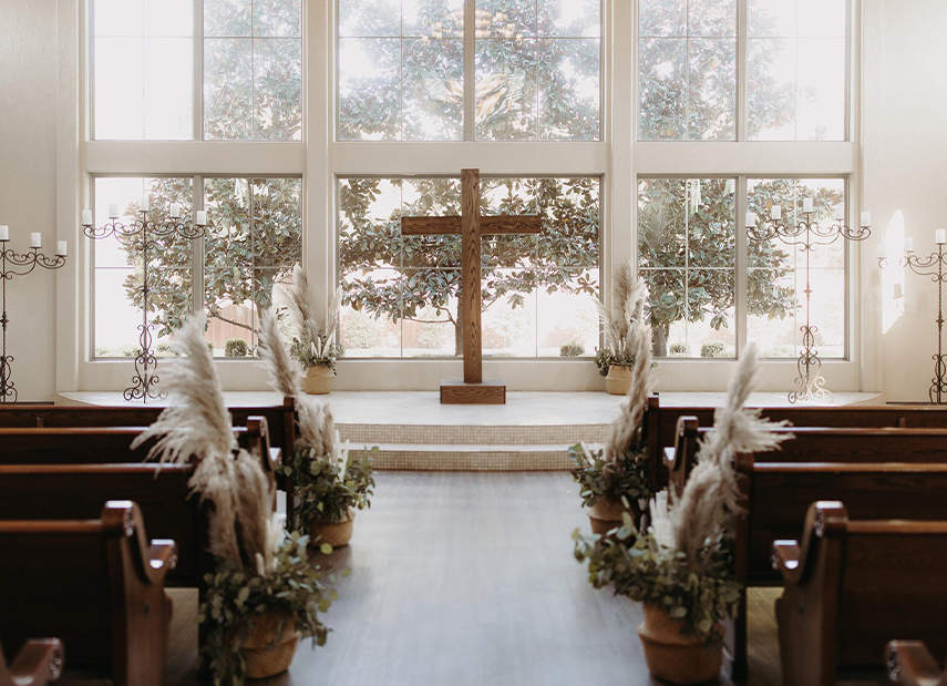 View up aisle of floor to ceiling windows with view of big tree, pews decorated with pampas grass
