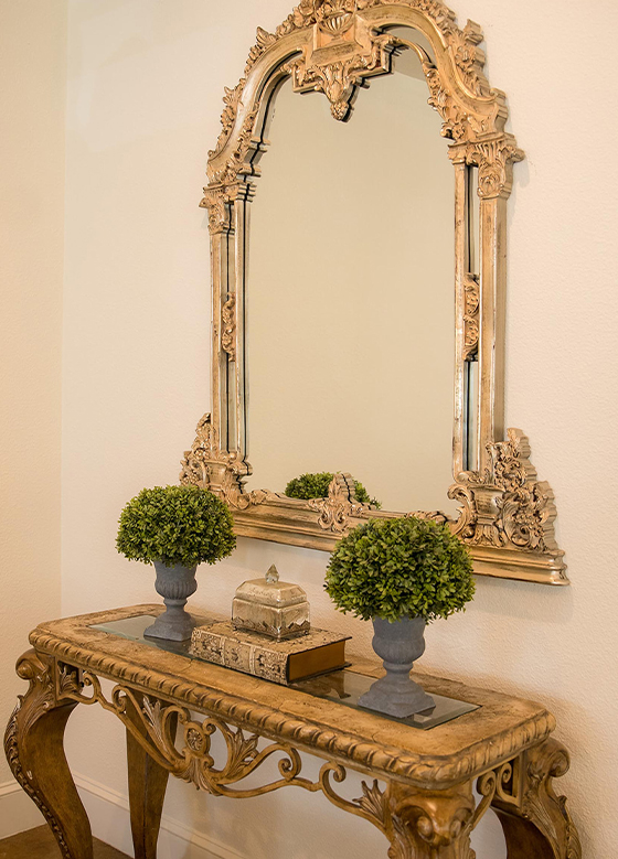 Side table with mini topiaries in front of gold ornate mirror