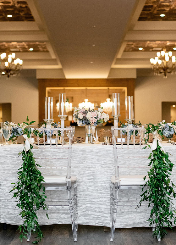 View of table in ballroom with clear chairs decorated with greenery