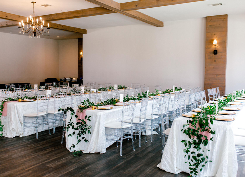 Banquet tables with clear chairs, white linens and greenery accents, white ceiling with wood beams and chandeliers