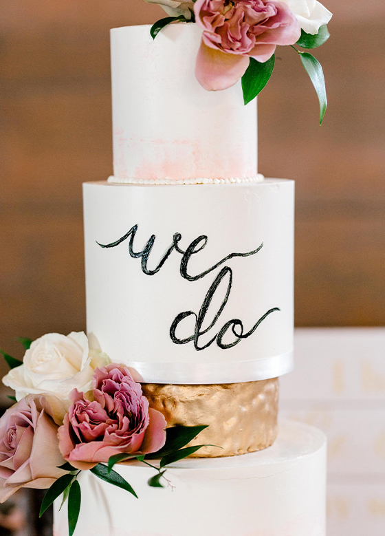 Close up of pretty white cake with "we do" in script and accent roses