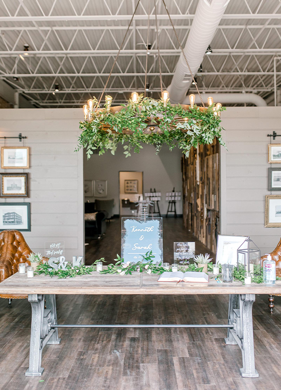 View of side table with greenery and keepsakes, a modern chandelier with Edison bulbs and greenery hangs above