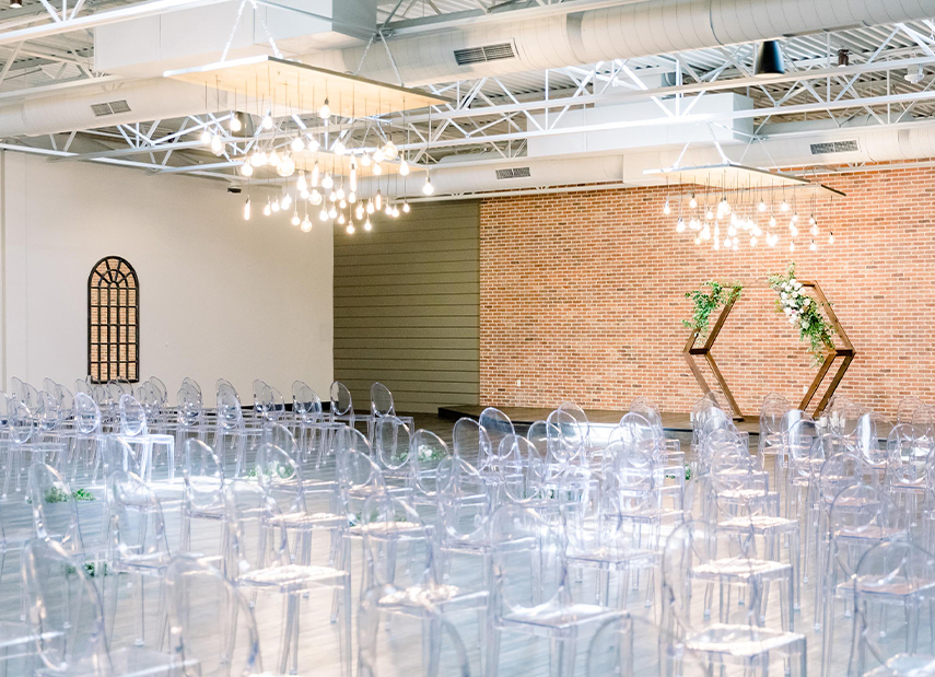 View across clear chairs towards ceremony set up with custom arch and brick wall
