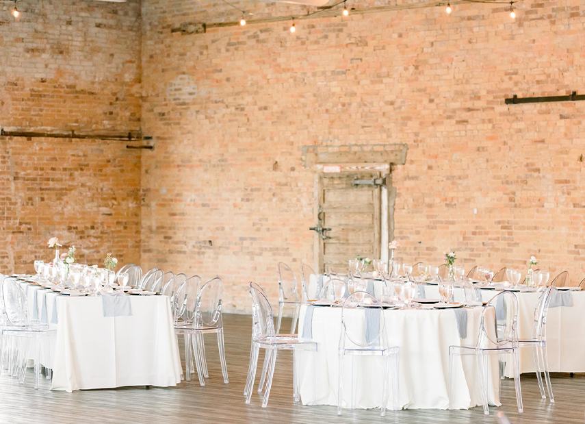 View of round tables with clear chairs and white linens against brick wall