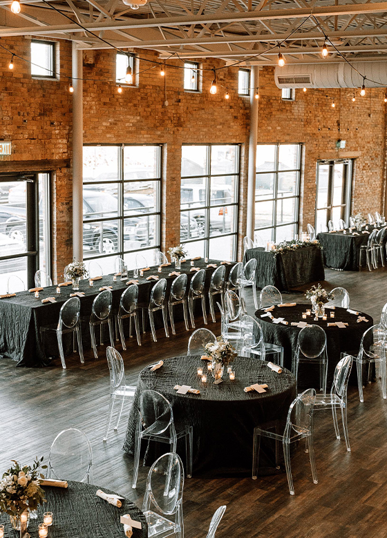 Wedding party banquet table and round tables with clear chairs and charcoal linens in brick industrial space