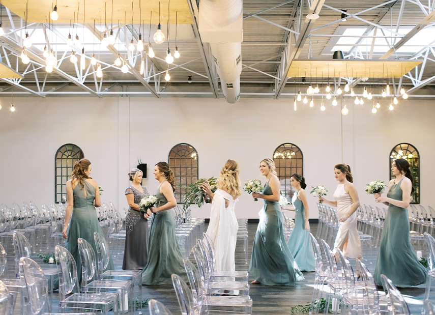 Bride and bridesmaids walking down aisle in industrial space with hanging lights and clear chairs
