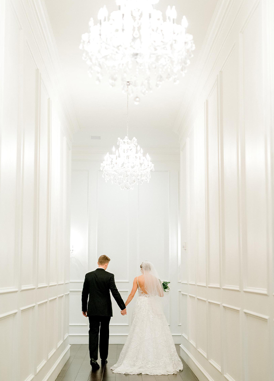Bride and groom walking through white hallway under chandeliers