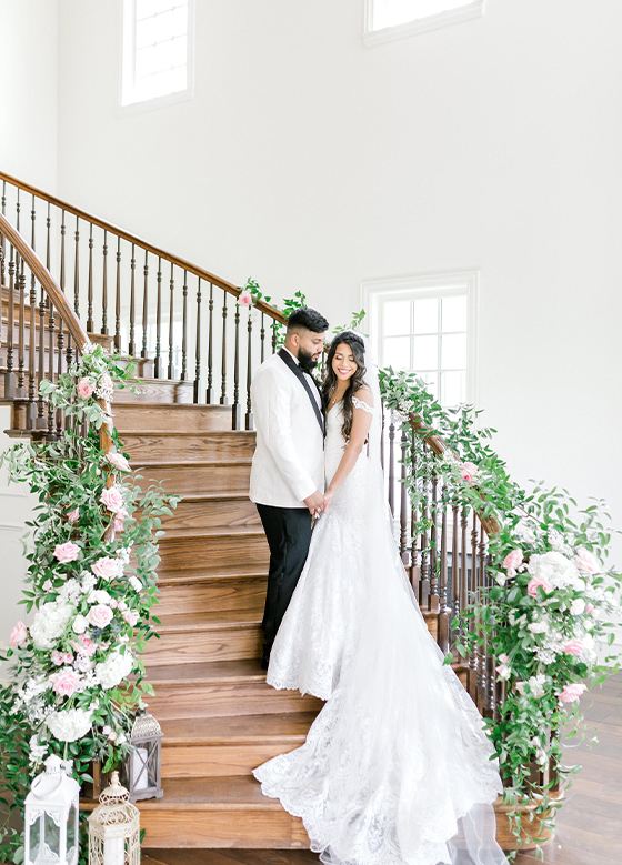 Bride and groom on grand wood staircase decorated with white florals and greenery