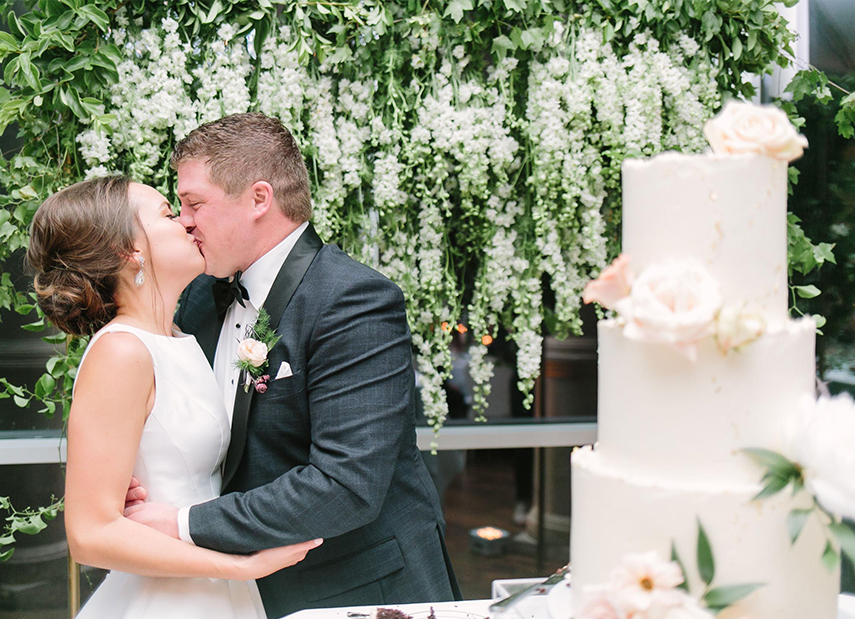 Bride and Groom Kiss Surrounded by Greenery Next to Simple Buttercream Wedding Cake