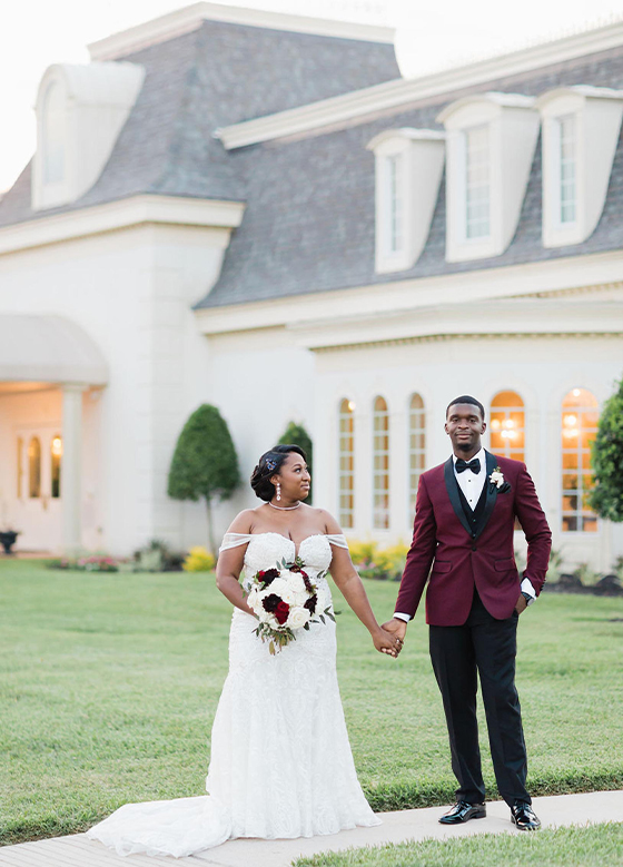 Groom in burgundy jacket holding hands with bride in white dress in front of wedding venue