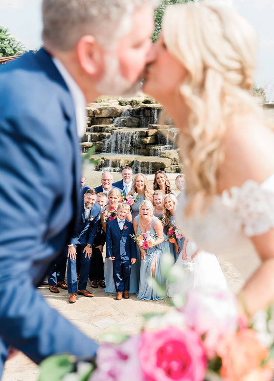 Out of Focus Bride Groom Kiss In Focus Wedding Party Behind with Bright Pink Floral Bouquets Fountain Greenery Rock Background